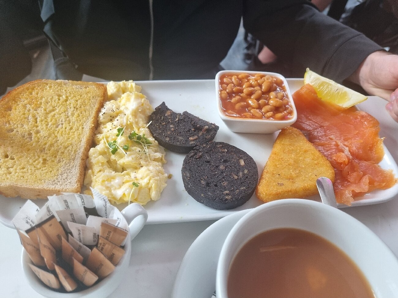 Full breakfast plate with toast, scrambled eggs, black pudding, baked beans, hash brown, and smoked salmon.