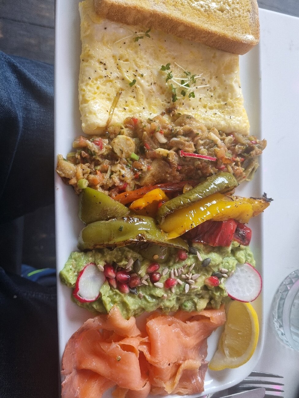 Plate with omelette, chilli aubergine, grilled peppers, smashed avocado, toast, and smoked salmon.