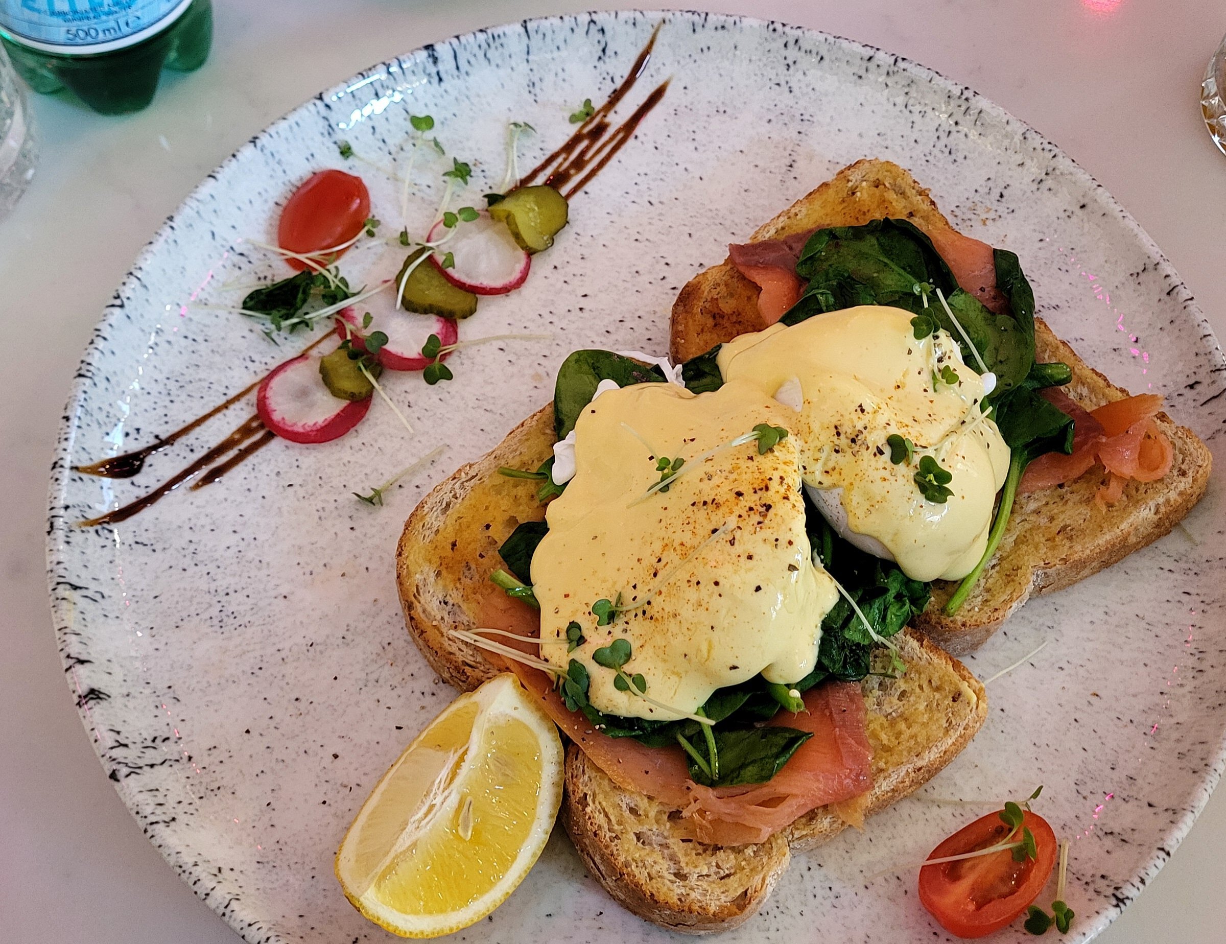 Two poached eggs with hollandaise, spinach, and smoked salmon on toast, garnished with lemon, radish, and microgreens.