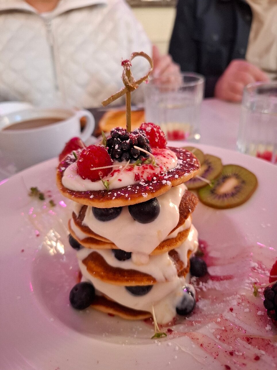 Stack of mini pancakes layered with yogurt, blueberries, and cream, topped with berries and powdered sugar.